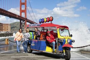 Three women smiling and posing next to a colorful red, blue, and yellow Lucky Tuk Tuk parked at Fort Point with the Golden Gate Bridge in the background.